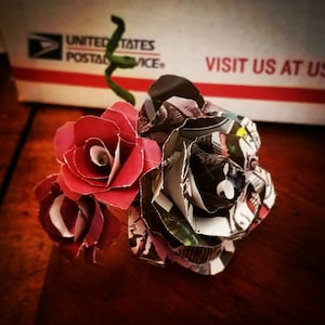 May include: A bouquet of three paper roses. Two roses are red and one is made from recycled comic book pages. The roses are arranged in a cluster.