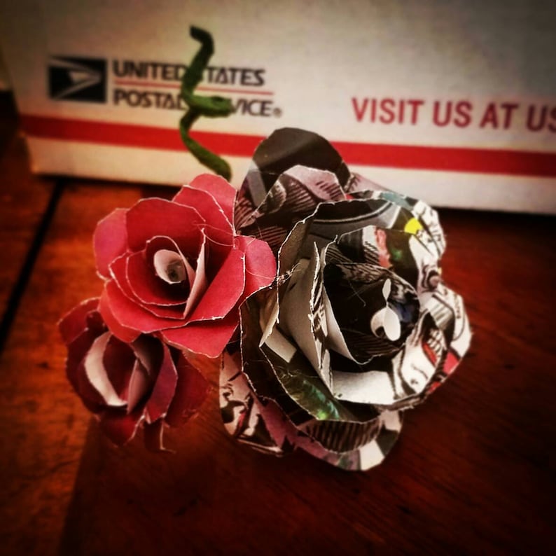 May include: A bouquet of three paper roses. Two roses are red and one is made from recycled comic book pages. The roses are arranged in a cluster.