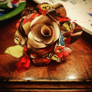 May include: A brown paper rose with colorful comic book paper petals. The rose is sitting on a wooden surface.