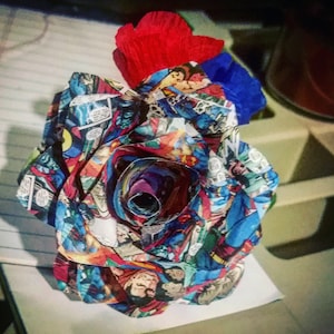 May include: A red, white, and blue paper rose made from comic book pages. The rose is curled into a spiral shape and has a red and blue paper bow on top.