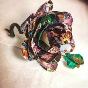 May include: A colorful paper flower made from comic book pages. The flower is a deep red, green, and blue with a black stem.