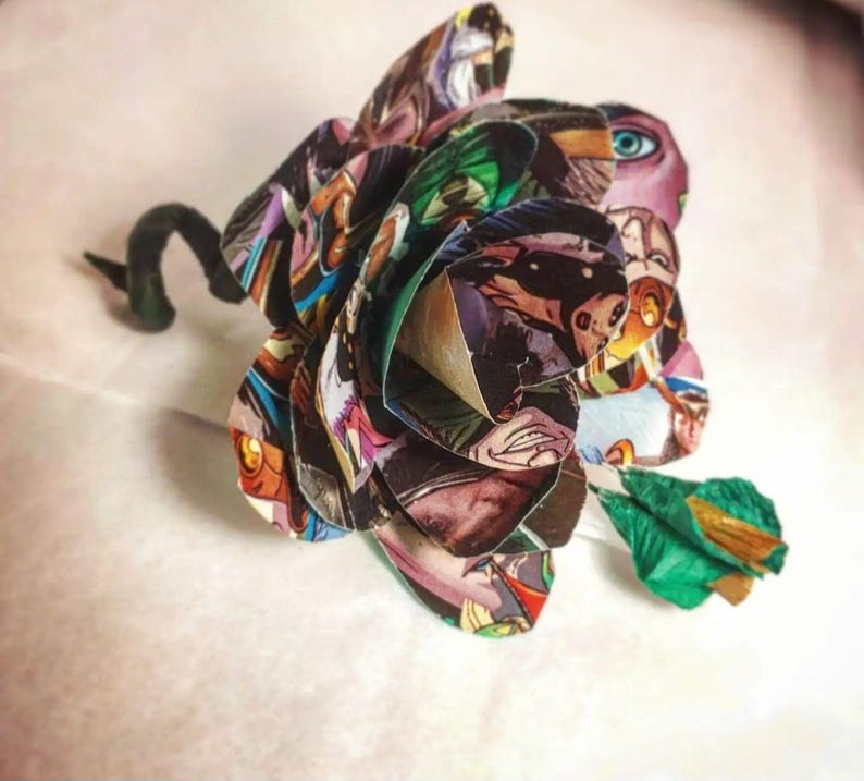 May include: A colorful paper flower made from comic book pages. The flower is a deep red, green, and blue with a black stem.