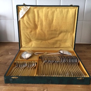 Puede incluir: Un juego de cubiertos antiguos de plata, presentado en una caja verde oscuro con interior amarillo. El juego incluye tenedores, cucharas y utensilios de servir. La cubertería plateada está cuidadosamente dispuesta dentro de la caja.