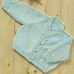 Könnte beinhalten: Hellblau gestrickte Baby-Strickjacke mit langen Ärmeln und Knopfleiste. Die Strickjacke hat ein Zopfmuster entlang der Knopfleiste und der Ärmelbündchen. Das Kleidungsstück ist aus weichem Garn gefertigt und liegt auf einer hellen Holzoberfläche.