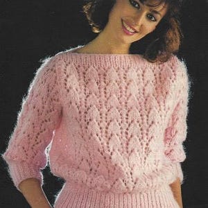 Può includere: Un maglione rosa chiaro, lavorato a maglia grossa spazzolata, con scollatura a barchetta e maniche a tre quarti. Il maglione presenta un motivo traforato strutturato e un orlo a coste. Il testo "BRUSHED CHUNKY" e "32-40 ins." (81-102 cm) è visibile.