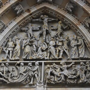 Pode incluir: Uma escultura em relevo de pedra detalhada que representa as Estações da Via Sacra. A escultura está localizada em uma porta arqueada e apresenta múltiplas figuras em diversas poses, incluindo uma figura central de Jesus Cristo na cruz.