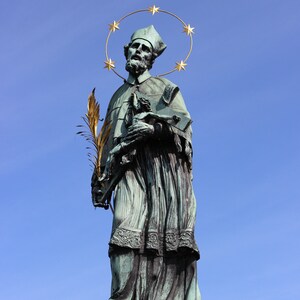 Pode incluir: Uma estátua de bronze de um homem vestindo uma túnica e uma auréola de estrelas. Ele está segurando uma pequena figura em seus braços. A estátua está em uma base de pedra.