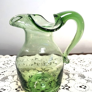 May include: A small green glass pitcher with a ruffled lip and a handle. The pitcher has a textured surface with small air bubbles.