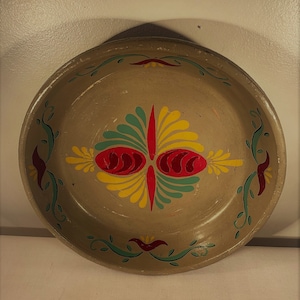 May include: A round, gray metal pie plate with a hand-painted design. The design features a central motif of stylized leaves and flowers in shades of red, yellow, and green.
