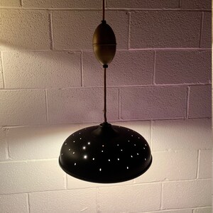 May include: A black metal pendant light with a perforated shade and a brass chain and canopy. The light is hanging from a white brick ceiling.