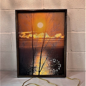 May include: A vintage sunset-themed clock with a brown wooden frame. The clock face features a black and white image of a sunset over a lake with trees in the foreground. The clock has a yellow cord.