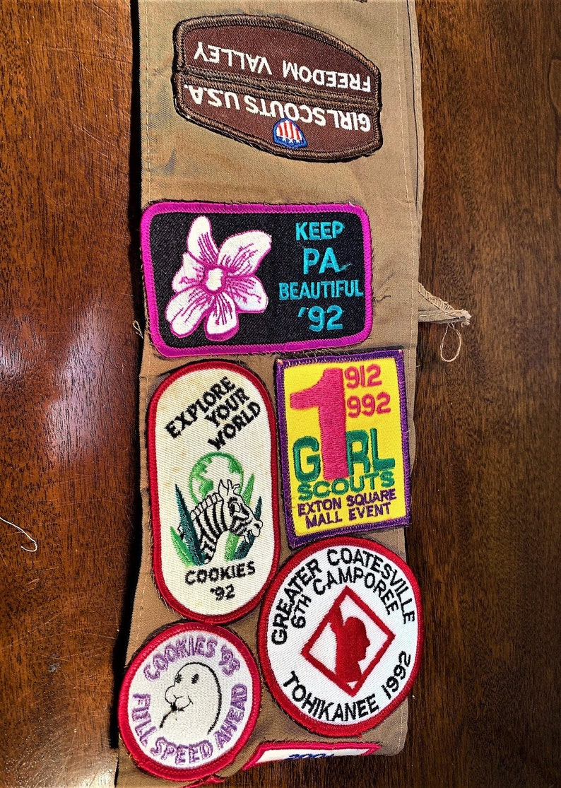 May include: A Girl Scout sash with various embroidered patches. The patches feature a flower, a zebra, a globe, a girl scout logo, and a camp logo. The text on the patches includes "Keep PA Beautiful '92", "Explore Your World", "Cookies '92", "1912 992 GRL Scouts Exton Square Mall Event", and "Greater Coatesville 6th Camporee Tohicanee 1992".