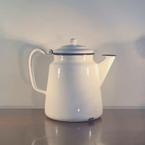 May include: White enamelware teapot with a black rim and handle. The teapot has a lid with a small knob and a spout for pouring. The handle is curved and attached to the side of the pot. The teapot is a classic kitchenware item.