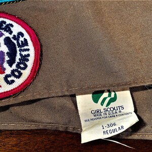 May include: A brown Girl Scouts sash with a green and white Girl Scouts logo patch and a white patch with a purple and red design that says "E6 COOKIES". The tag on the sash says "GIRL SCOUTS MADE IN U.S.A. SEE REVERSE FOR CARE & CONTENTS 1-306 REGULAR".