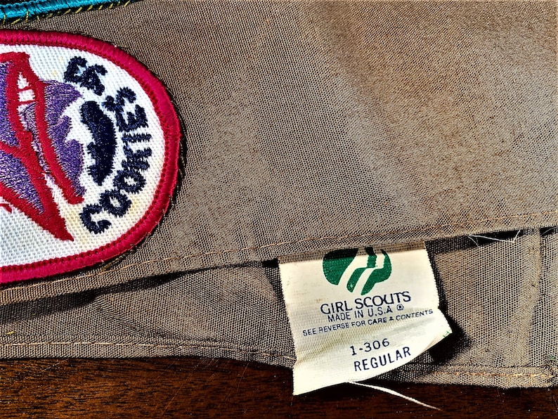 May include: A brown Girl Scouts sash with a green and white Girl Scouts logo patch and a white patch with a purple and red design that says "E6 COOKIES". The tag on the sash says "GIRL SCOUTS MADE IN U.S.A. SEE REVERSE FOR CARE & CONTENTS 1-306 REGULAR".