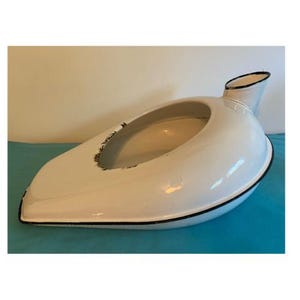 May include: A white, vintage bedpan with a black trim. The bedpan has an oval opening and a spout. The bedpan is made of a glossy material and sits on a blue surface.