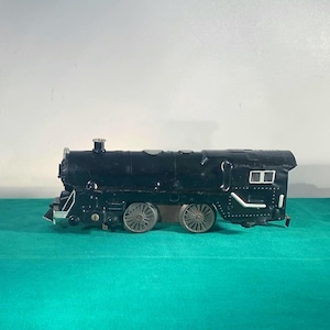 May include: A black toy train with silver wheels and accents. The train has a detailed design, including a smokestack, headlights, and windows. It is set against a green surface and a white background.