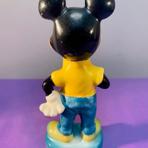 Pu&ograve; includere: Una statuetta in ceramica di Topolino con una camicia gialla e jeans blu. &Egrave; in piedi su una base blu con un bordo bianco.