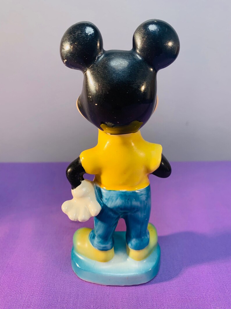 Pu&ograve; includere: Una statuetta in ceramica di Topolino con una camicia gialla e jeans blu. &Egrave; in piedi su una base blu con un bordo bianco.