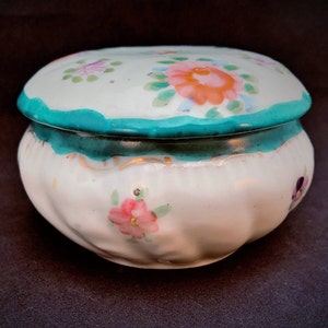 May include: A white ceramic trinket box with a lid. The lid and body are decorated with pink, orange, and purple floral designs. The rim of the lid and the base of the box are trimmed with teal paint.