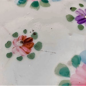 May include: Close-up of a white surface, possibly ceramic, adorned with hand-painted floral patterns. The flowers feature pink, red, purple, and blue hues, complemented by green leaf accents. The design has a delicate, artistic quality.
