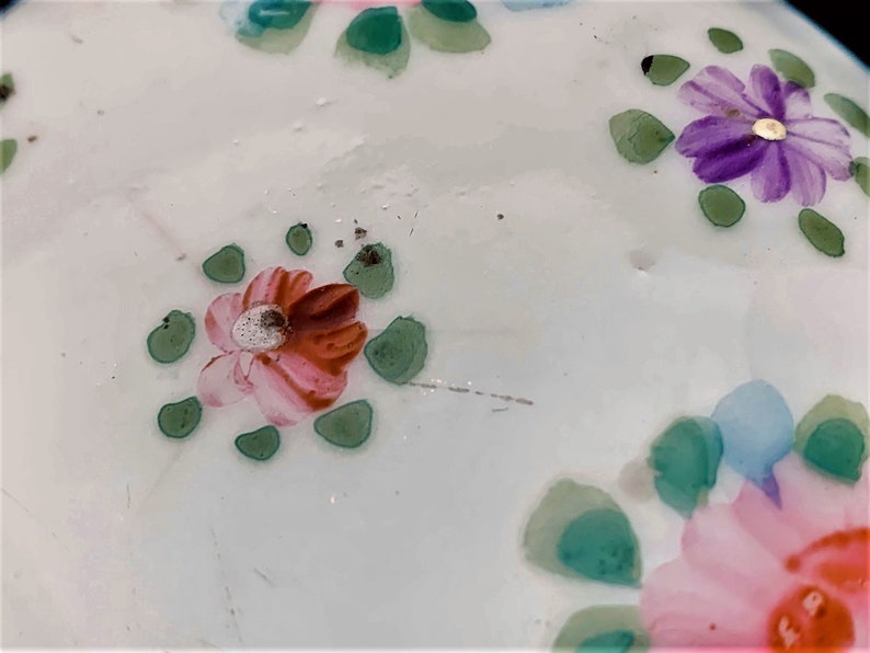 May include: Close-up of a white surface, possibly ceramic, adorned with hand-painted floral patterns. The flowers feature pink, red, purple, and blue hues, complemented by green leaf accents. The design has a delicate, artistic quality.