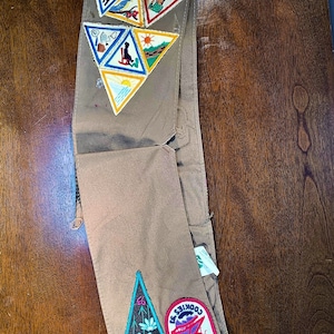 May include: A brown Girl Scout sash with various embroidered patches, including a triangular patch with the text "Keep PA Beautiful" and a rectangular patch with the text "Full Speed Ahead Cookies '63".