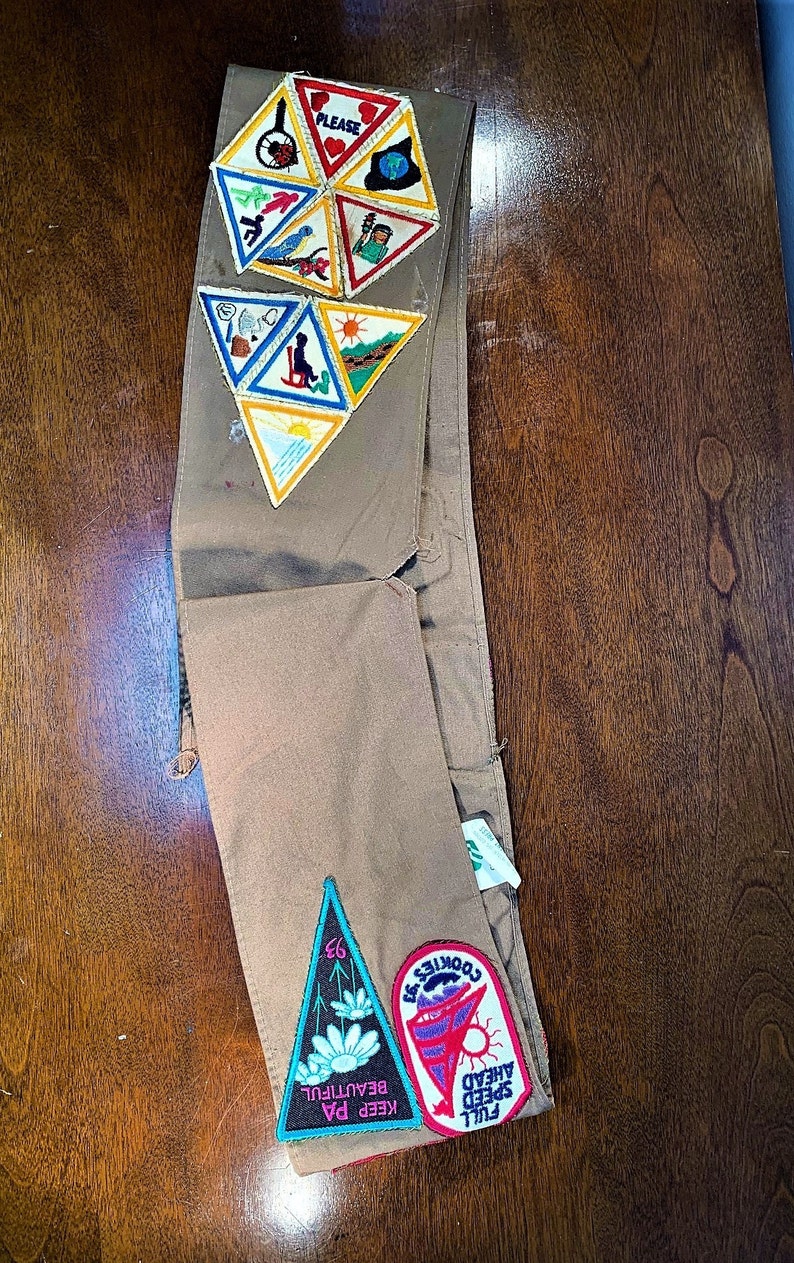 May include: A brown Girl Scout sash with various embroidered patches, including a triangular patch with the text "Keep PA Beautiful" and a rectangular patch with the text "Full Speed Ahead Cookies '63".