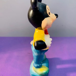 Pu&ograve; includere: Una statuetta in ceramica di Topolino con una camicia gialla, jeans blu e un papillon rosso. &Egrave; in piedi su una base blu con accenti gialli.