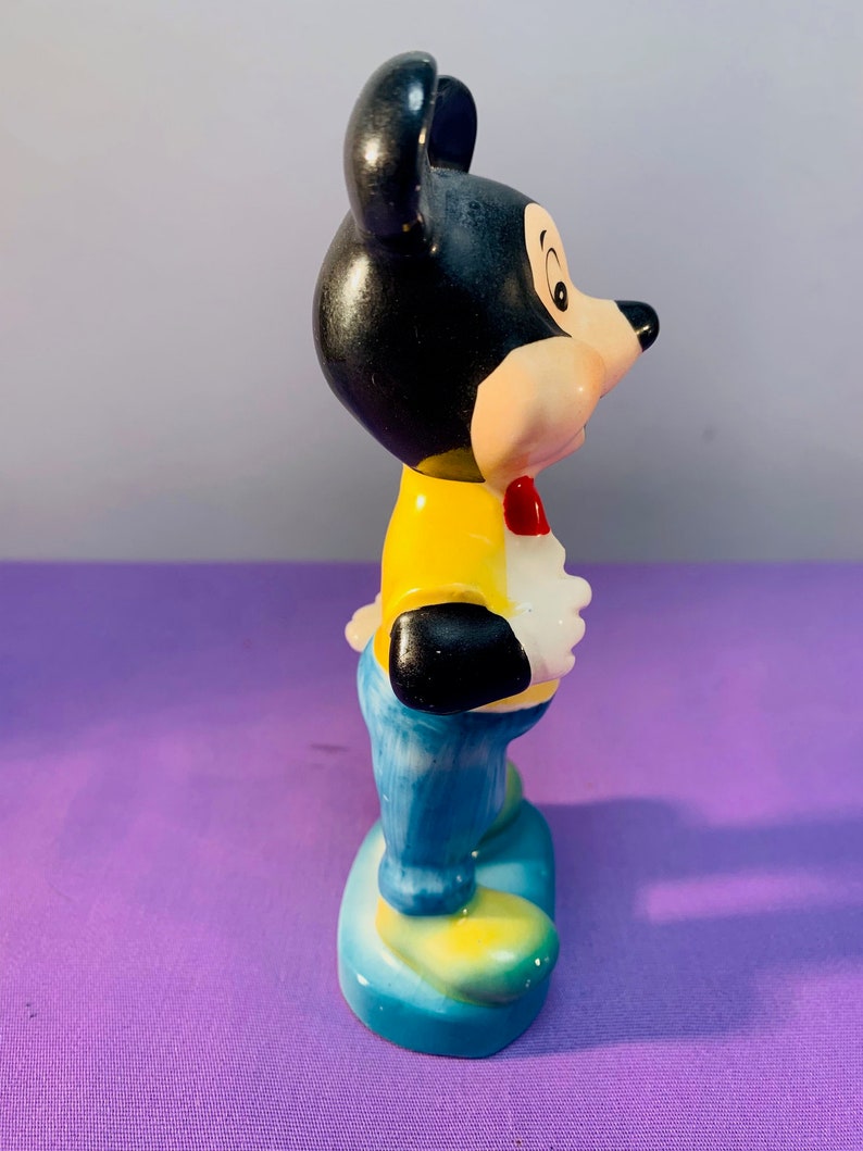 Pu&ograve; includere: Una statuetta in ceramica di Topolino con una camicia gialla, jeans blu e un papillon rosso. &Egrave; in piedi su una base blu con accenti gialli.