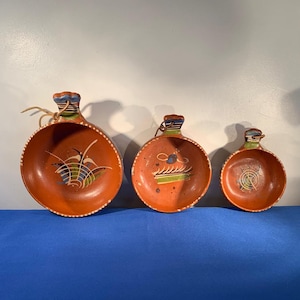 May include: Three terracotta bowls with handles, each painted with different colorful designs. The bowls are nested together, with the largest bowl in the back and the smallest bowl in the front.