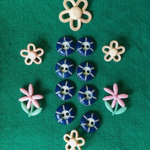 May include: An assortment of sewing buttons in various shapes and colors. The image features several navy blue buttons with white detailing, along with flower-shaped buttons in cream and pink. The buttons are arranged on a green fabric background.