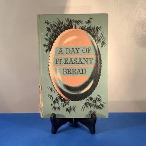 May include: Vintage cookbook titled "A Day of Pleasant Bread" on a black stand. The cover is light blue with an oval design in peach and black, and the title is in white. The book is slightly worn.