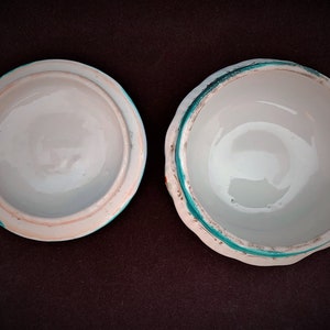 May include: An open white ceramic trinket box with a lid. The lid and the box have a light blue rim with a turquoise accent. The interior of both pieces is white. The box is displayed on a dark brown surface.