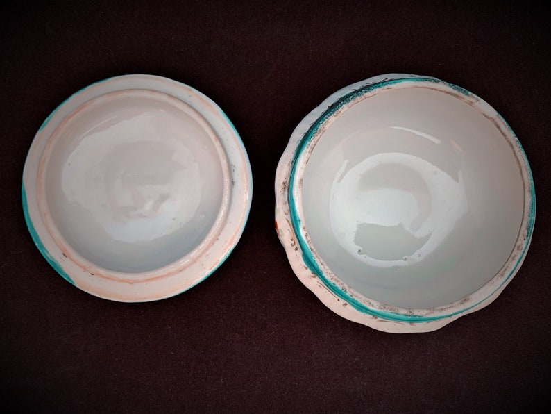 May include: An open white ceramic trinket box with a lid. The lid and the box have a light blue rim with a turquoise accent. The interior of both pieces is white. The box is displayed on a dark brown surface.