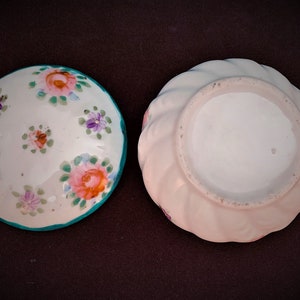 May include: Two small, round ceramic dishes. One is white with pink, orange, and purple floral designs and a teal rim. The other is a light pink with a swirled edge.