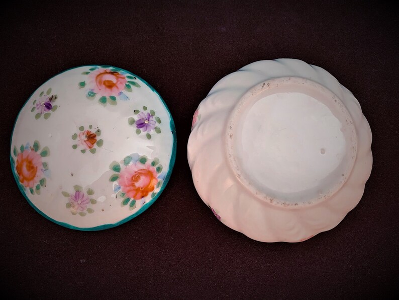 May include: Two small, round ceramic dishes. One is white with pink, orange, and purple floral designs and a teal rim. The other is a light pink with a swirled edge.