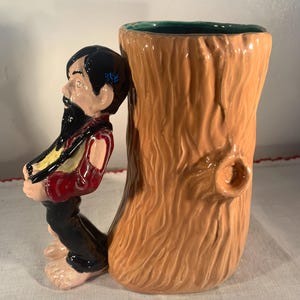 May include: A whimsical ceramic mug featuring a bearded man figure leaning against a tree trunk. The mug has a brown exterior with a green interior. The man is dressed in a red vest, black pants, and has black hair. The mug is a unique decorative item.