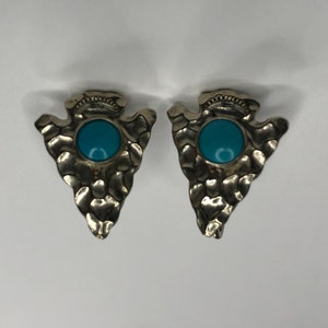 May include: A pair of silver-toned arrowhead earrings with a textured surface. Each earring features a round, turquoise-colored stone in the center. The earrings are displayed against a white background.