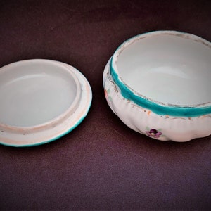 May include: A white ceramic trinket box with a lid, featuring a turquoise rim and floral details. The box has a scalloped edge and a white interior. The lid is separate, showing the inside of the box. The box is open.