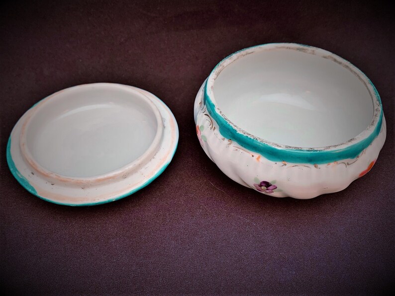 May include: A white ceramic trinket box with a lid, featuring a turquoise rim and floral details. The box has a scalloped edge and a white interior. The lid is separate, showing the inside of the box. The box is open.