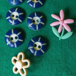 May include: A collection of sewing buttons in various designs. There are several navy blue, circular buttons with white accents, and two flower-shaped buttons. One flower is pink with green leaves, and the other is a cream color.