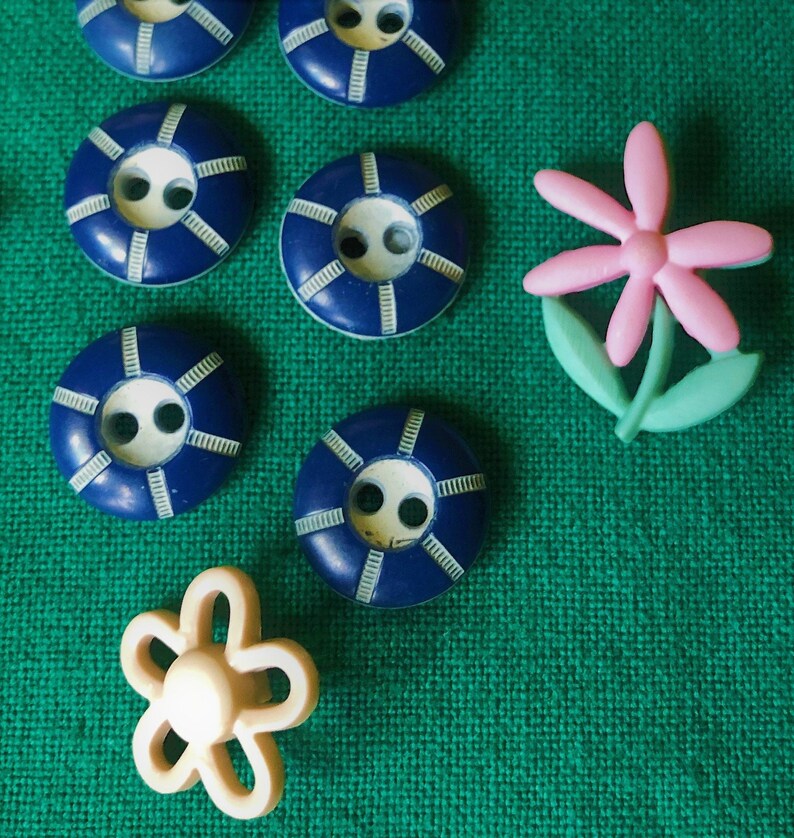 May include: A collection of sewing buttons in various designs. There are several navy blue, circular buttons with white accents, and two flower-shaped buttons. One flower is pink with green leaves, and the other is a cream color.