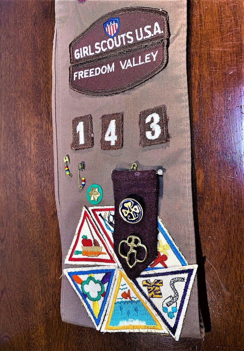 May include: A Girl Scouts USA sash with the words "Freedom Valley" and the number "143" on it. The sash has several badges and pins, including a green four-leaf clover, a brown patch with a four-leaf clover, and a brown patch with a red apple and a boat.
