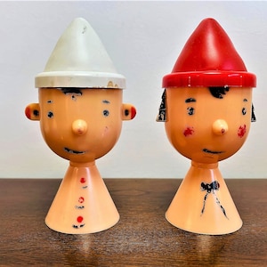 May include: Two vintage ceramic egg holders shaped like people with painted faces. One has a white hat and the other has a red hat.