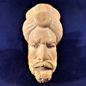 May include: A white ceramic pipe shaped like a man's head wearing a turban. The pipe has a detailed face with a beard and mustache.