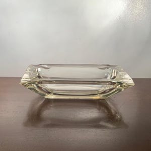 May include: A clear, rectangular glass ashtray with a slightly beveled edge. The ashtray has a shallow bowl and is designed with a classic, minimalist aesthetic. The glass reflects light, creating a subtle play of shadows.