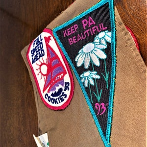 May include: A brown Girl Scout vest with two embroidered patches. The left patch is blue, pink, and white with the text "Full Speed Ahead" and "Cookies". The right patch is black, white, pink, and teal with the text "Keep PA Beautiful" and "93".