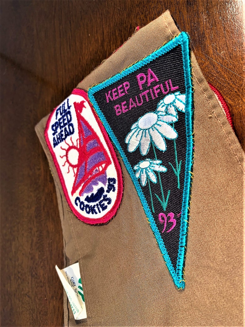 May include: A brown Girl Scout vest with two embroidered patches. The left patch is blue, pink, and white with the text "Full Speed Ahead" and "Cookies". The right patch is black, white, pink, and teal with the text "Keep PA Beautiful" and "93".
