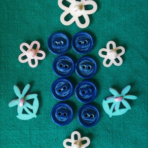 May include: An assortment of sewing buttons and embellishments on a green background. The collection includes blue round buttons, white and pink flower-shaped buttons, and turquoise butterfly-shaped embellishments.